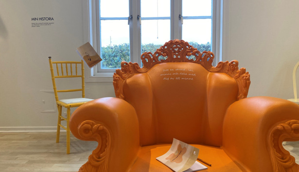 En orange stol med en bok på toppen av den. Min Historia utställning.