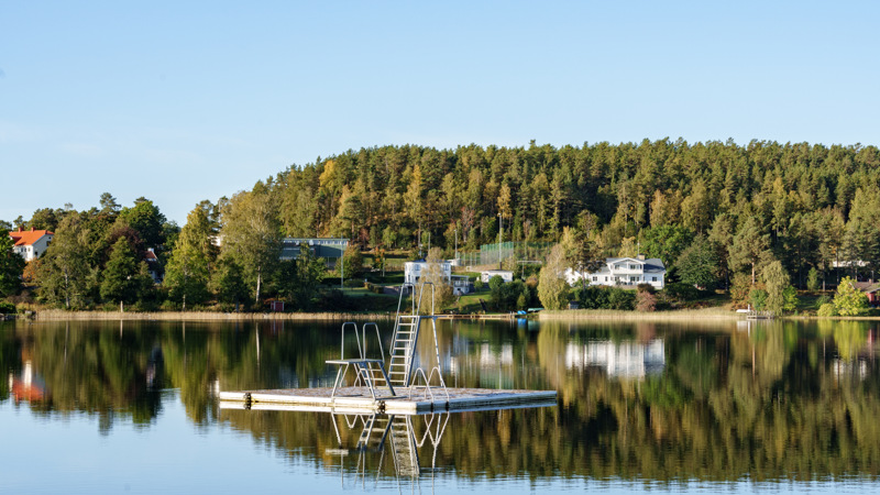Ett hopptorn på flotte i vattnet med träd och hus i bakgrunden