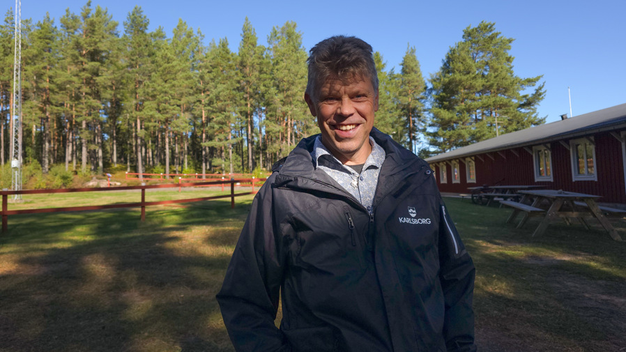 Man står på en öppen gräsyta med ett hus i bakgrunden samt en mindre skogsdunge.