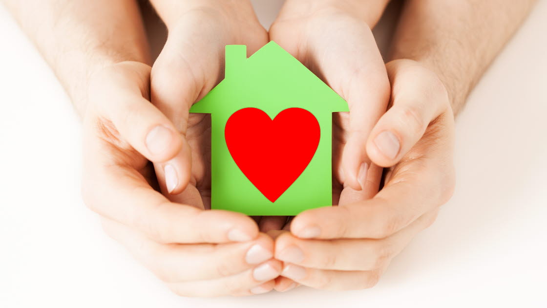 Händer håller i ett grönt litet hus med ett rött hjärta i.